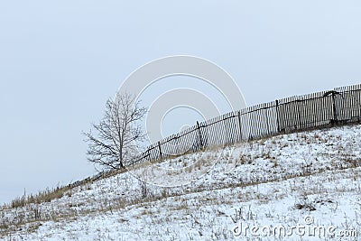 A sad landscape against a gray sky, snow and a crooked fence Stock Photo