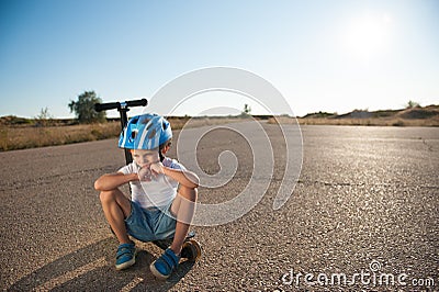 Sad little active kid in sport protective helmet sitting lonely on scooter on empty summer asphalt highway on hot sunset with copy Stock Photo