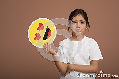 Sad little girl holding face with heart eyes emoji on pale pink background Stock Photo