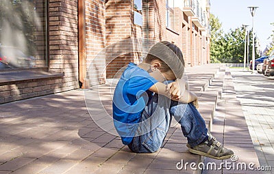 Sad, lonely, unhappy, disappointed child sitting alone on the ground. City background. Outdoor Stock Photo