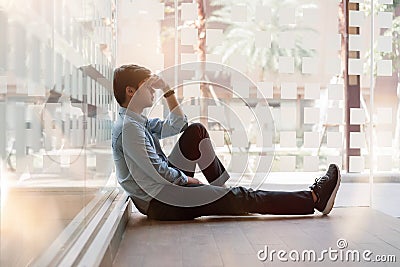 Sad man sitting on the floor by window at office walkway feeling stressed holding head with hands. Depression and anxiety disorder Stock Photo