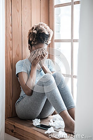 Darkskinned woman covering face with hands sitting on windowsill Stock Photo