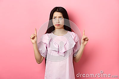 Sad pitiful asian girl sulking, pointing and looking up with disappointed face, feeling upset, standing against pink Stock Photo