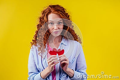 Sad redhaired ginger curly cry female tearing up red broken paper heart in studio yellow background..sex education Stock Photo