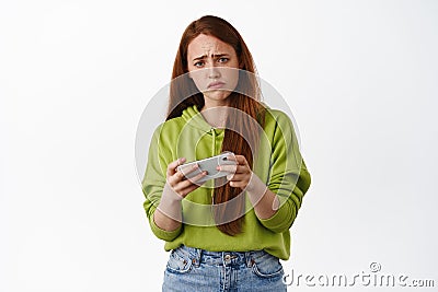 Sad redhead girl losing video game on mobile phone, watching upsetting video on smartphone and grimacing, frowning Stock Photo