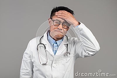 Sad sick funny adult asian man doctor in white coat, glasses touches forehead, suffers from fever Stock Photo