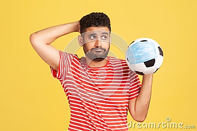 Sad thoughtful man in red t-shirt holding soccer ball in protective mask, symbol of football competition during covid-19 virus, Stock Photo