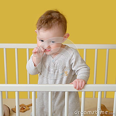 Sad toddler baby boy in the crib gnawing a nail, yellow studio background. A tired child in pajamas Stock Photo