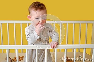 Sad toddler baby boy in the crib gnawing a nail, yellow studio background. A tired child in pajamas Stock Photo