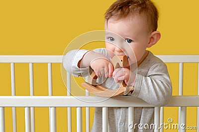 Sad toddler baby boy in the crib gnawing a wooden toy, yellow studio background. A tired child in pajamas Stock Photo