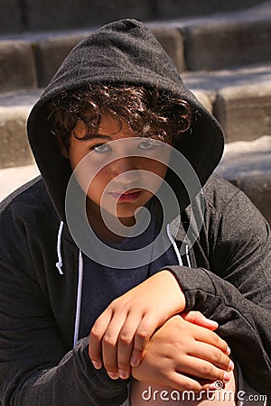 Sad troubled teenager school boy with hood on posing outdoor sitting alone on the street Stock Photo