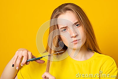 Sad and unhappy teen girl cutting her hair with scissors while standing. Young student experiments with her hairstyle at home Stock Photo