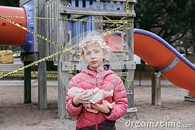 Sad upset Caucasian girl with baby toy on closed playground outdoor. Kids play area locked with yellow caution tape in Toronto, Stock Photo