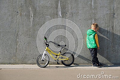 Sad upset little boy standing near concrete wall with his bicycle on sunny day. Urban lifestyle Stock Photo