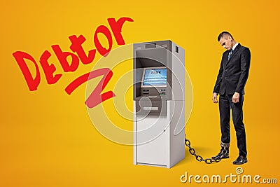 Sad young businessman standing in half-turn chained to ATM on amber background with big red title `Debtor`. Stock Photo