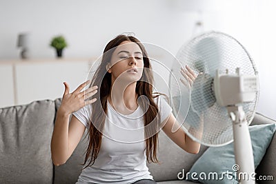 Sad young european woman suffers from unbearably too hot weather, catches cold air from fan in living room Stock Photo