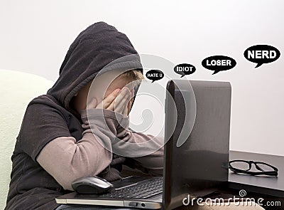 A sad boy in a hoodie, with his face in his hands, is sitting at a table, in front of a laptop, alone. Insults fly Stock Photo