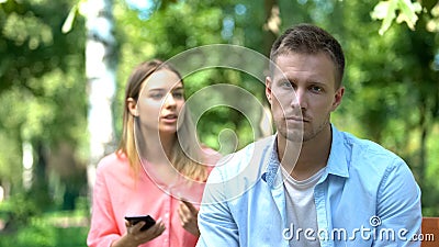 Sad young man looking camera, annoyed jealous girlfriend holding phone, distrust Stock Photo