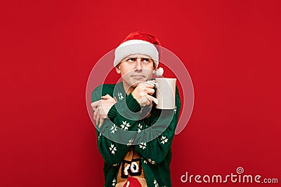 Sad young man in a Santa hat and Christmas sweater warms with a cup of tea in his hands and looks away on red background. Guy in Stock Photo