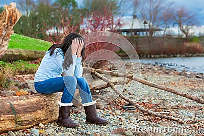 Sad young teen girl sitting on log along rocky beach by lake Stock Photo