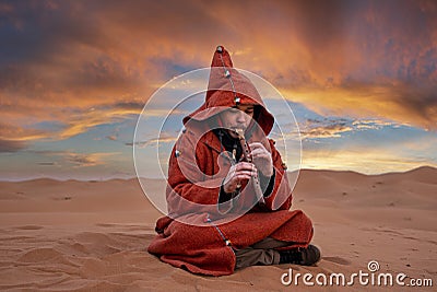 Man in traditional red clothes playing flute while sitting on sand in desert Editorial Stock Photo