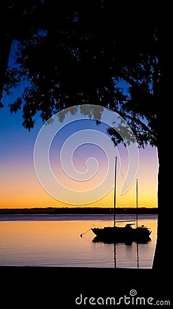A sailboat moored at sunset viewed through frame of a tree silhouette against a colorful sky - room for copy Stock Photo