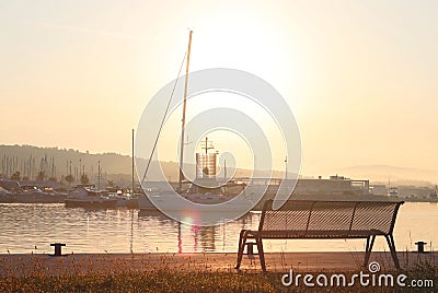 The sailing yacht enters under the motor in the marina in the rays of the morning sun. Dawn in the port. Empty bench on the pier Stock Photo