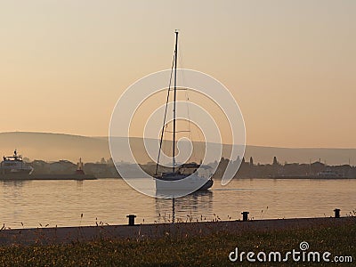 The sailing yacht enters under the motor in the marina in the rays of the morning sun. Dawn in the port. Empty pier with electrica Stock Photo