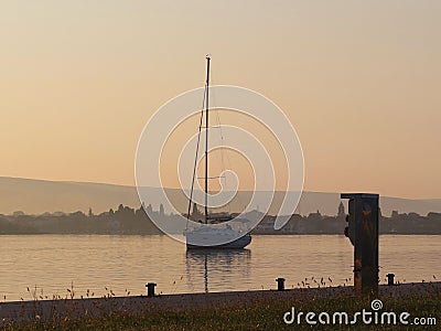 The sailing yacht enters under the motor in the marina in the rays of the morning sun. Dawn in the port. Empty pier with Stock Photo