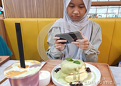 Samarinda, Indonesia. January 23th 2023. Happy Asian muslim teenage girl sitting in cafe and taking picture of her food with smart Stock Photo