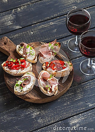 Sandwiches with goat cheese, anchovies, roasted peppers, ham and two glasses of red wine on a wooden rustic board. Stock Photo