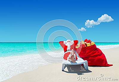 Santa Claus with Christmas sack full of gifts relax on sunlounger barefooted at perfect sandy ocean beach. Stock Photo