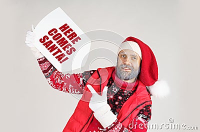 Santa Claus holds a sign with the inscription in his hands - HERE COMES SANTA and points to it with his other hand Stock Photo