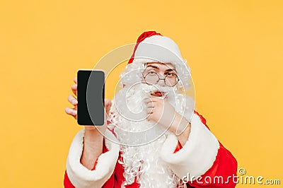 Santa Claus stands on a yellow background, looks at the camera with a pensive face and shows a smartphone with a black screen. Stock Photo