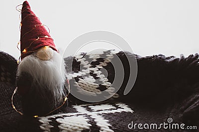 Santa Claus toy with garland on grey knitted background. Cute dwarf with red conical hat in scandinavian design. Stock Photo