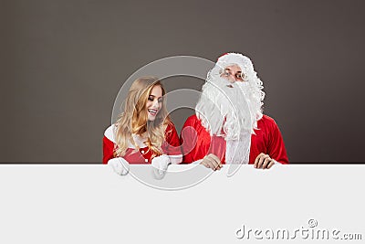 Santa Claus and young beautiful mrs. Claus stand behind a white canvas and smile on the gray background. Stock Photo