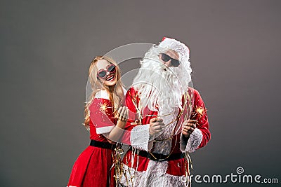 Santa Claus and young beautiful mrs. Claus in sunglasses are holding sparklers in their hands on the gray background Stock Photo