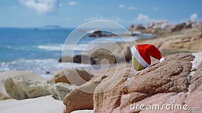 Santa Hat on Coconut on tropical sandy beach with ocean waves rolling to rocky coastline in blurred background. stock footage Santa Hat on Coconut on tropical sandy beach with ocean waves rolling to rocky coastline in blurred background. stock footage
