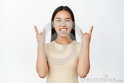 Sassy brunette asian girl shows heavy metal, rock on gesture, sticking tongue and looking excited, having fun, standing Stock Photo