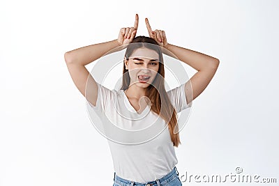 Sassy and flirty young woman wink, shows tongue and bull devil horns gesture, fingers on head, being assertive and Stock Photo