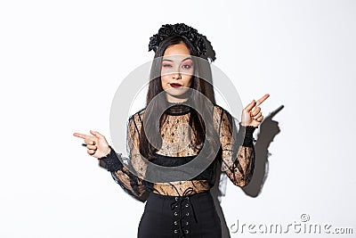 Sassy young evil witch with gothic makeup and wreath, looking arrogant while pointing fingers sideways, showing two Stock Photo