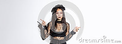 Sassy young evil witch with gothic makeup and wreath, looking arrogant while pointing fingers sideways, showing two Stock Photo