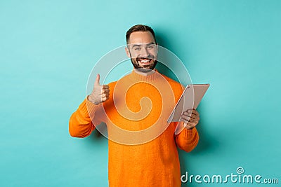 Satisfied adult man smiling, using digital tablet and showing thumb-up, approve and agree, standing against turquoise Stock Photo