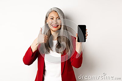 Satisfied asian elderly businesswoman showing blank smartphone screen and thumb-up, praising online promotion or company Stock Photo