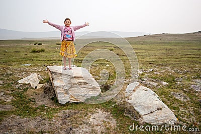 Satisfied child girl stands with arms spread out on a stone on a field with green grass. Stock Photo