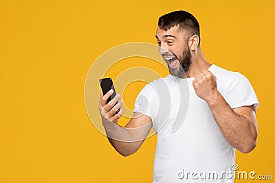 Satisfied excited adult european guy in white t-shirt looking at smartphone and expresses joy, celebrating success Stock Photo