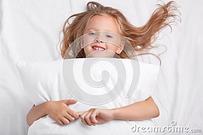 Satisfied playful girl with charming smile, embraces pillow, lies on white pillow, has good rest, enjoys awakening, poses in bed. Stock Photo