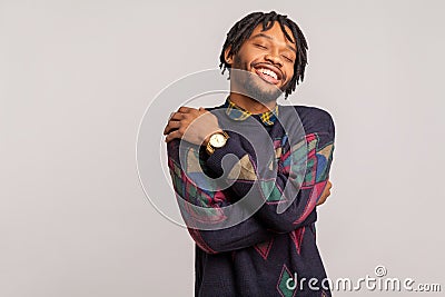 Satisfied pleased african man in casual sweatshirt with dreadlocks hugging himself, egoistically comforting and relaxing, self Stock Photo