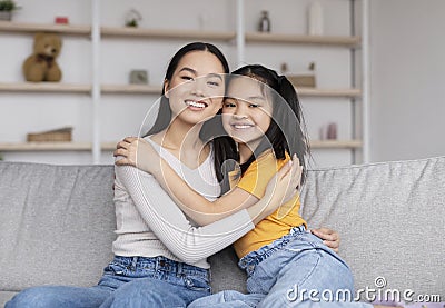 Satisfied pretty millennial chinese woman and teenage girl hugging, sitting on sofa in living room Stock Photo