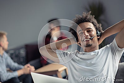 Satisfied relaxed african-american freelancer feeling happy at work sitting at office desk with headset. Stock Photo